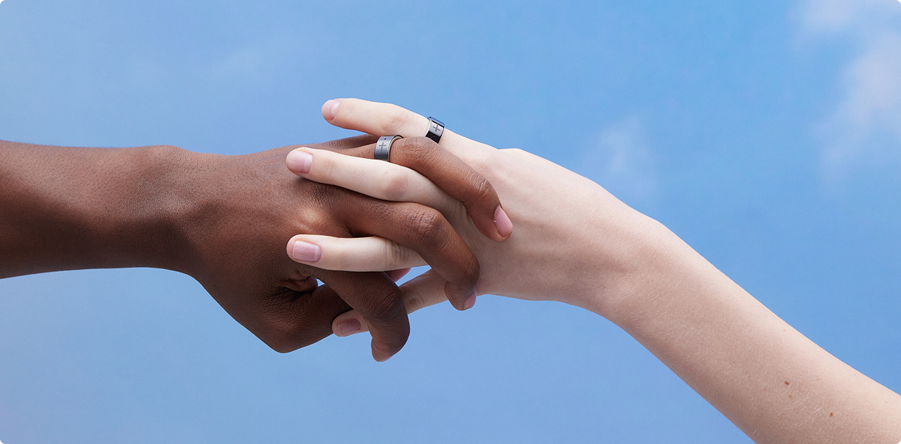 Two hands clasped together, wearing Bless Rings against a blue sky
